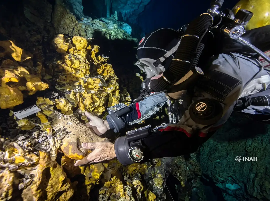 A scuba diver in full gear examines yellow-encrusted rock formations underwater inside a cave system