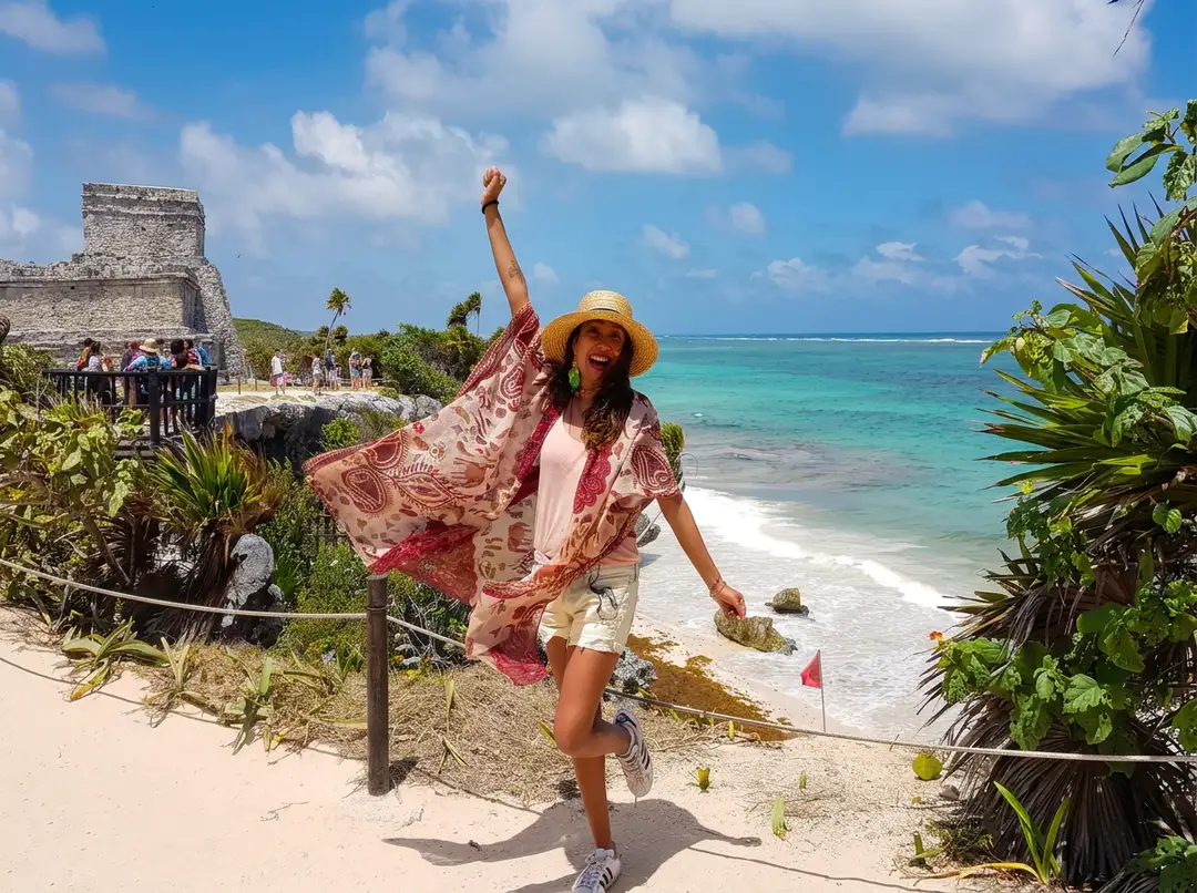 Woman in a straw hat and flowing pink cover-up poses with her fist raised on a sandy beach with turquoise water, an ancient stone structure, and tropical vegeta