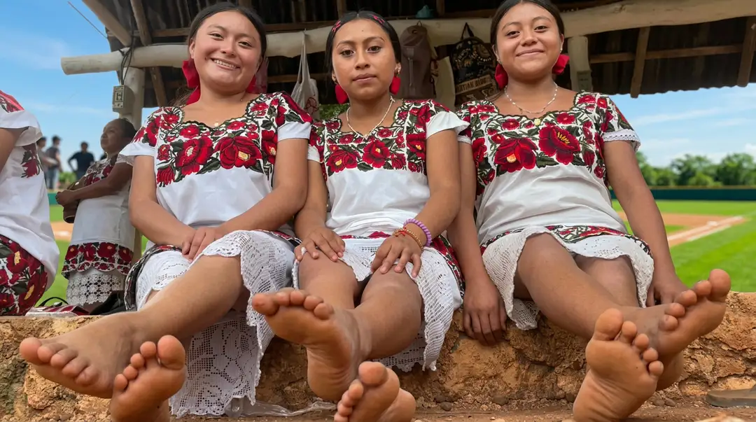 Three young women in matching embroidered red and white traditional dresses sit together on the ground near a baseball field