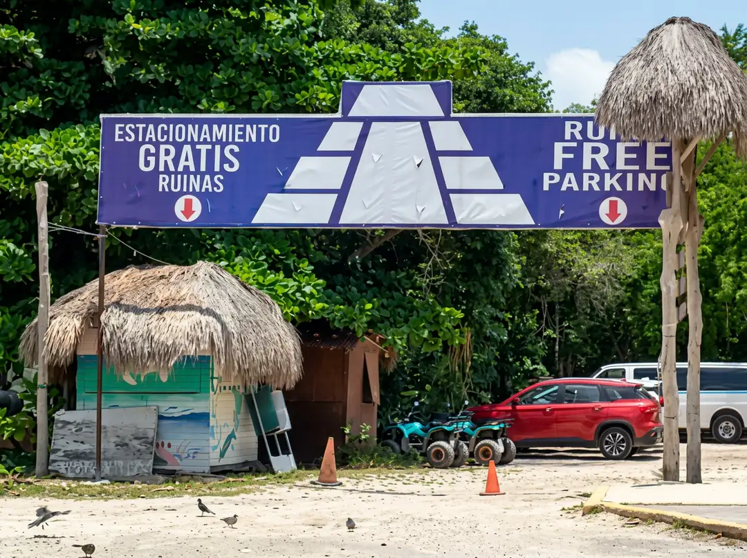 A blue and white parking sign with thatched-roof structures and parked vehicles beneath dense green vegetation near a sandy lot