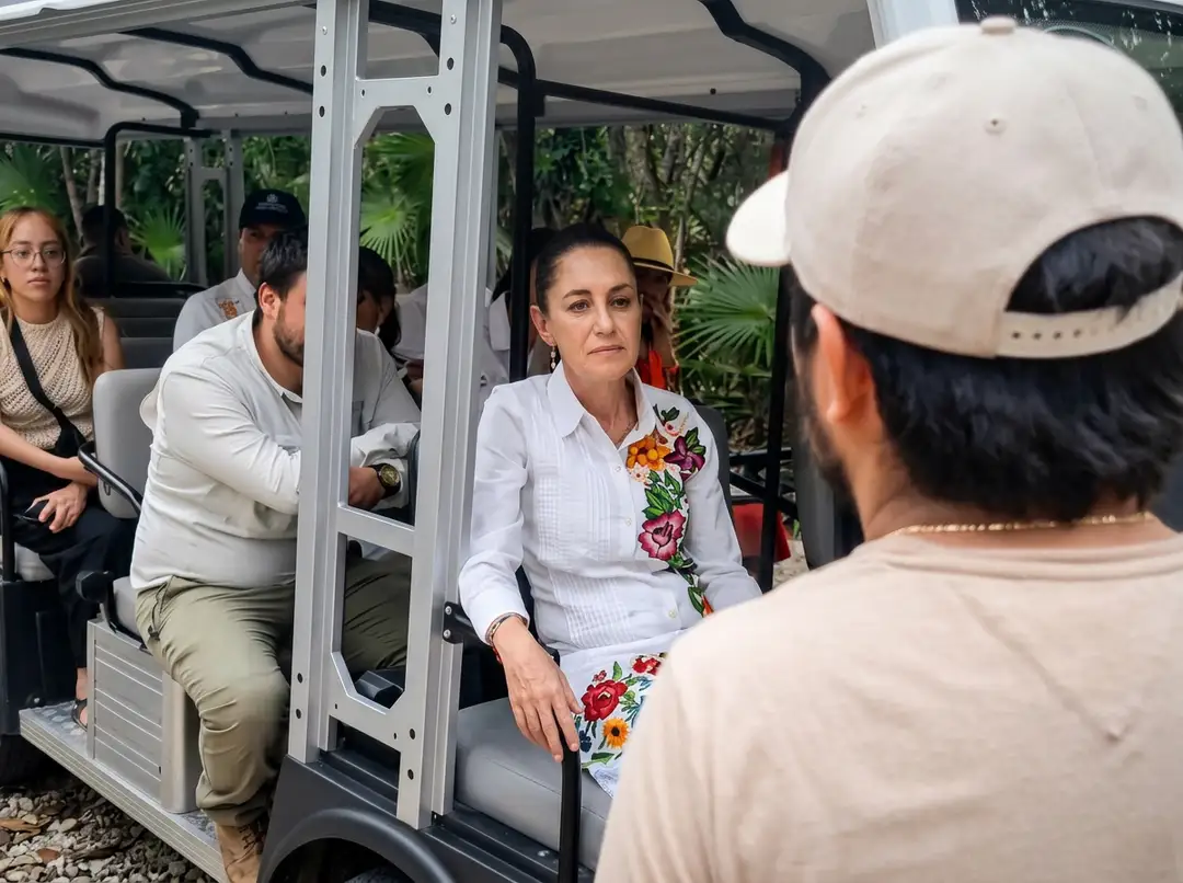 A woman in a white shirt with a floral design sits in an open-air tram car surrounded by tourists and lush greenery, speaking with a man wearing a white cap out
