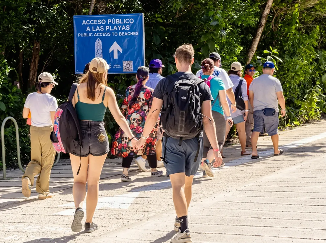 Should Tulum Remain Open Year-Round? A group of tourists walks along a shaded path toward a blue sign indicating public beach access, with tropical vegetation on either side