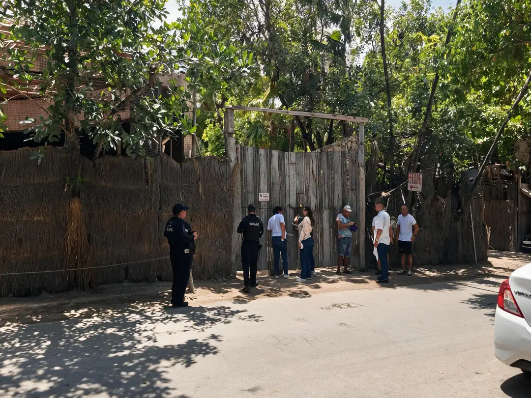 Judge Orders Possession Returned in Tulum Land Dispute and Clears Officials to Break Locks Police officers stand outside a wooden gate while a group of people wait in line on a sandy street shaded by trees