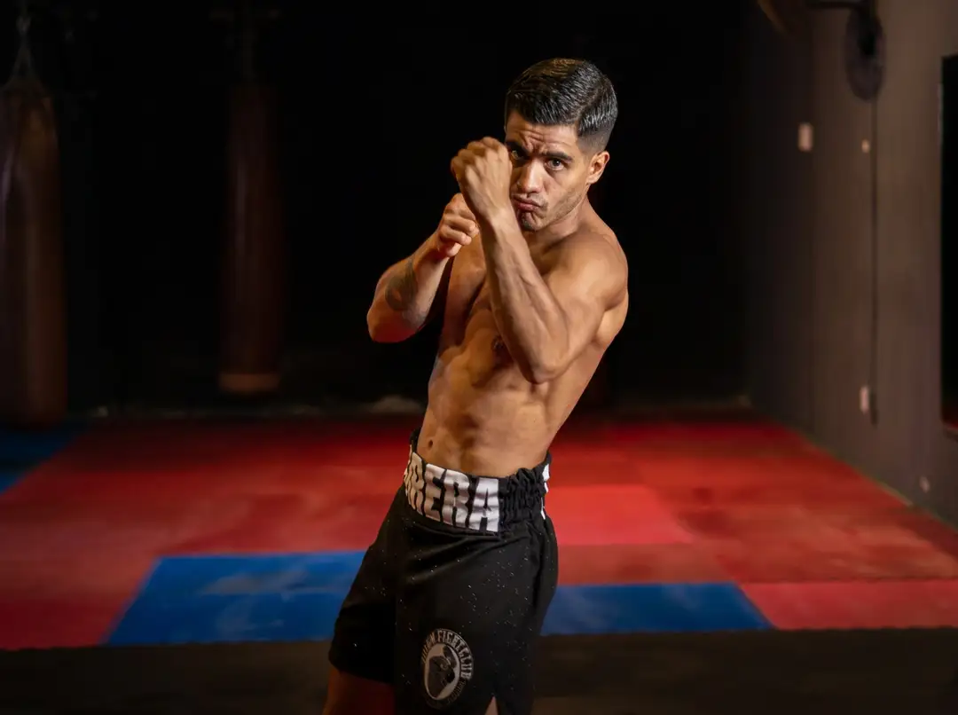 Shirtless fighter in boxing stance on red and blue mat in darkened gymnasium