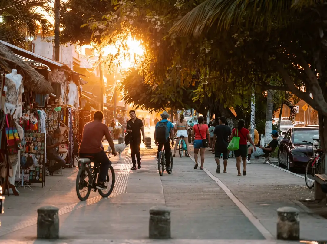 People bike and walk along a tree-lined pedestrian street with vendor stalls and shops during sunset