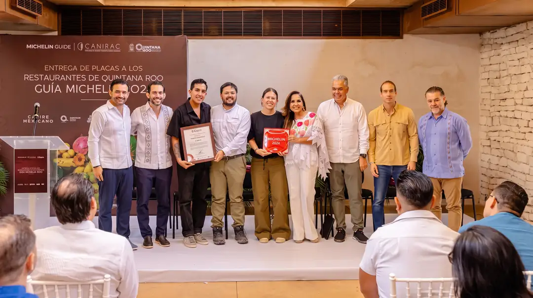 Michelin named eight restaurants in Tulum. The state named the milpa and the market Nine people stand on a stage holding certificates and awards at a restaurant recognition ceremony, with an audience seated in the foreground