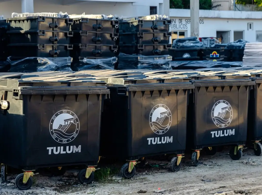 Large black waste containers marked Tulum sit parked in a lot with other industrial bins and vehicles visible in the background