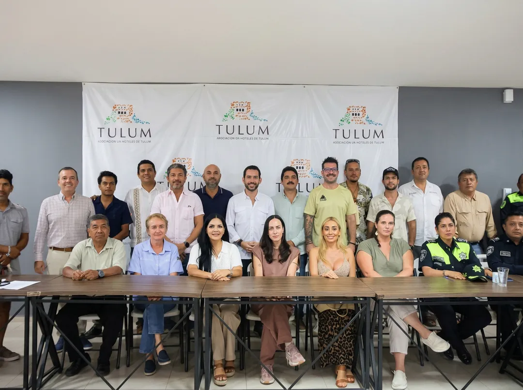 Large group of people seated and standing behind a table at a press conference or public meeting with Tulum banners in the background