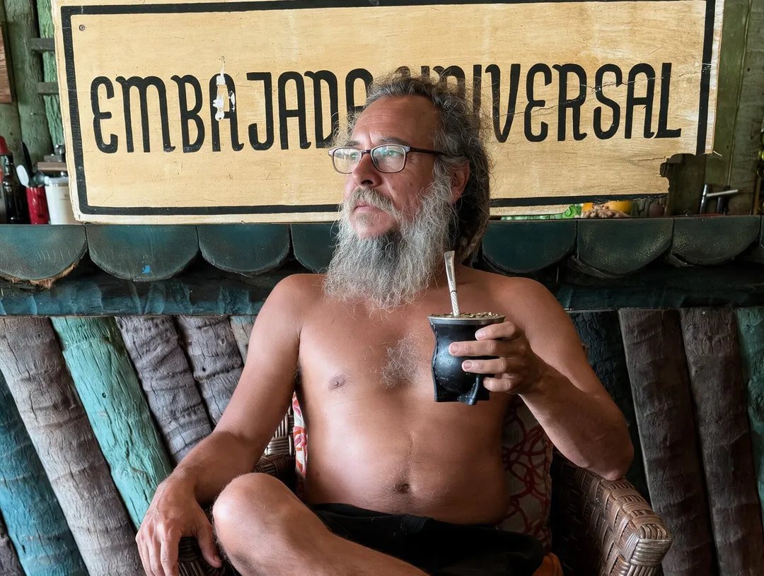 Man with gray beard and glasses sits shirtless holding a drink in front of a wooden sign reading "Embajador Universal