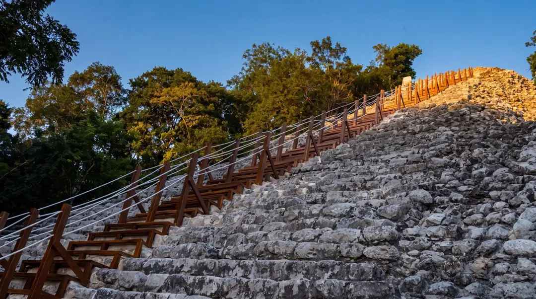 Wooden staircase with handrail runs diagonally up a stone pyramid structure surrounded by trees under clear sky