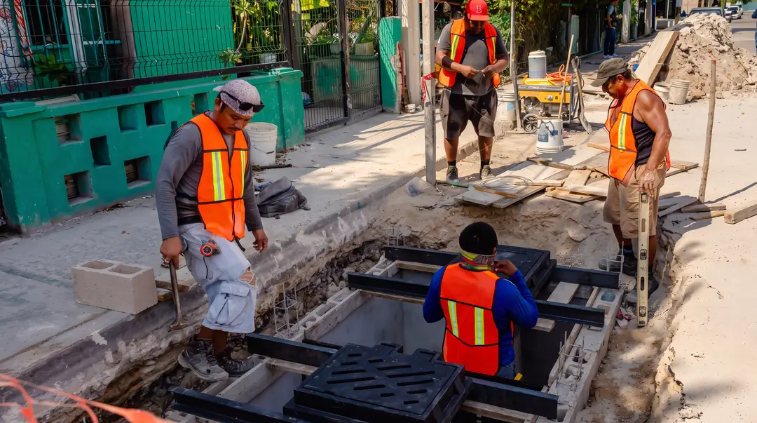 Construction workers in safety vests install drainage grates into an open trench on a city street lined with green buildings