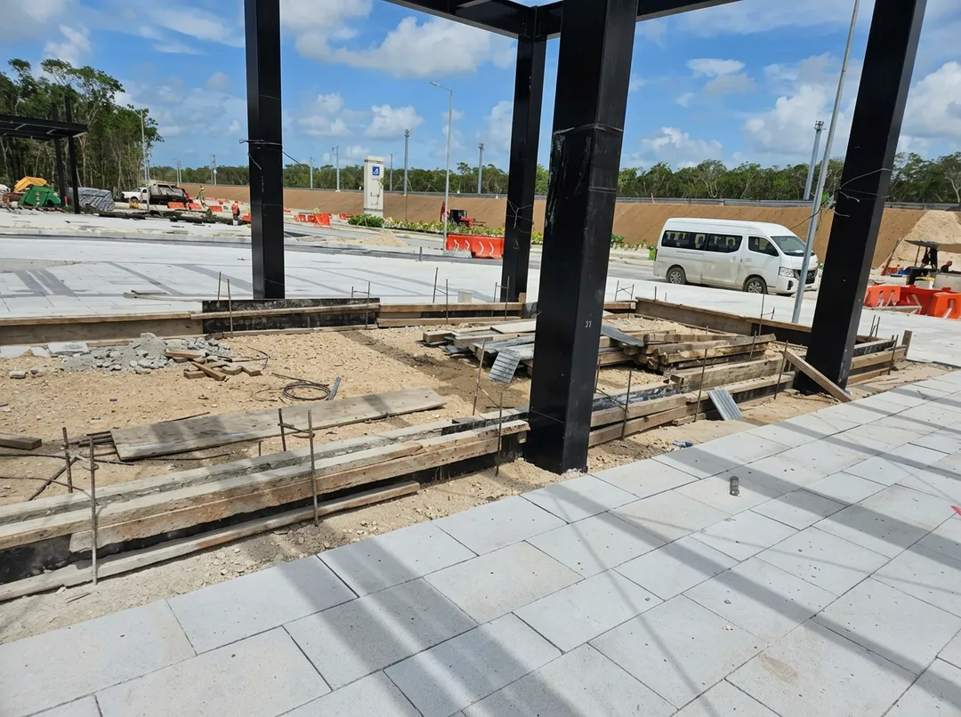 Construction site with concrete foundation work and large black support columns under a steel structure, with vehicles and equipment visible in an open area wit