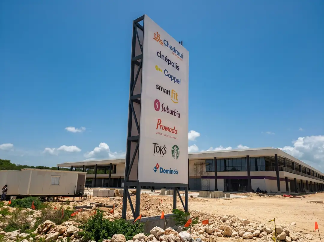 A tall directional sign listing multiple retail brand names stands in front of an under-construction commercial building against a clear blue sky
