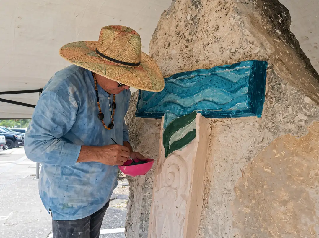 Artist in straw hat and blue shirt paints a letter on a stone wall with natural pigments