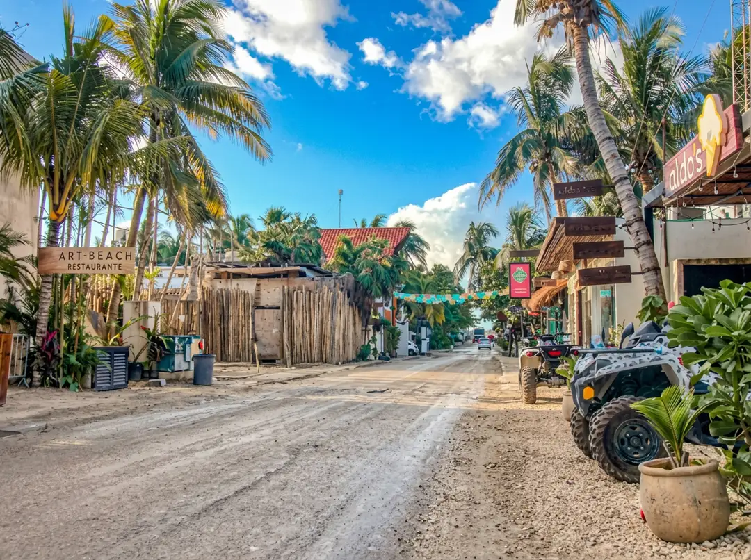 A sandy street lined with palm trees and commercial buildings, with an all-terrain vehicle parked on the right and few people visible on a clear day