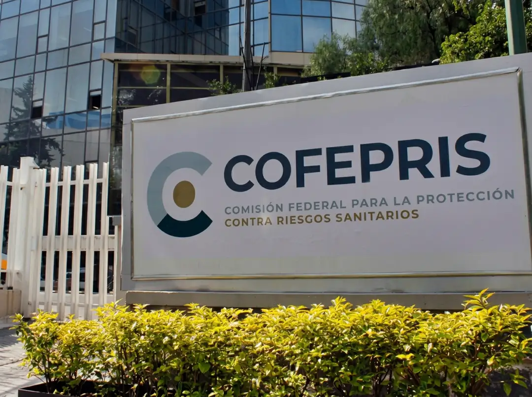 A sign for Cofepris, a federal health protection agency, stands in front of a modern glass building with yellow shrubs at its base
