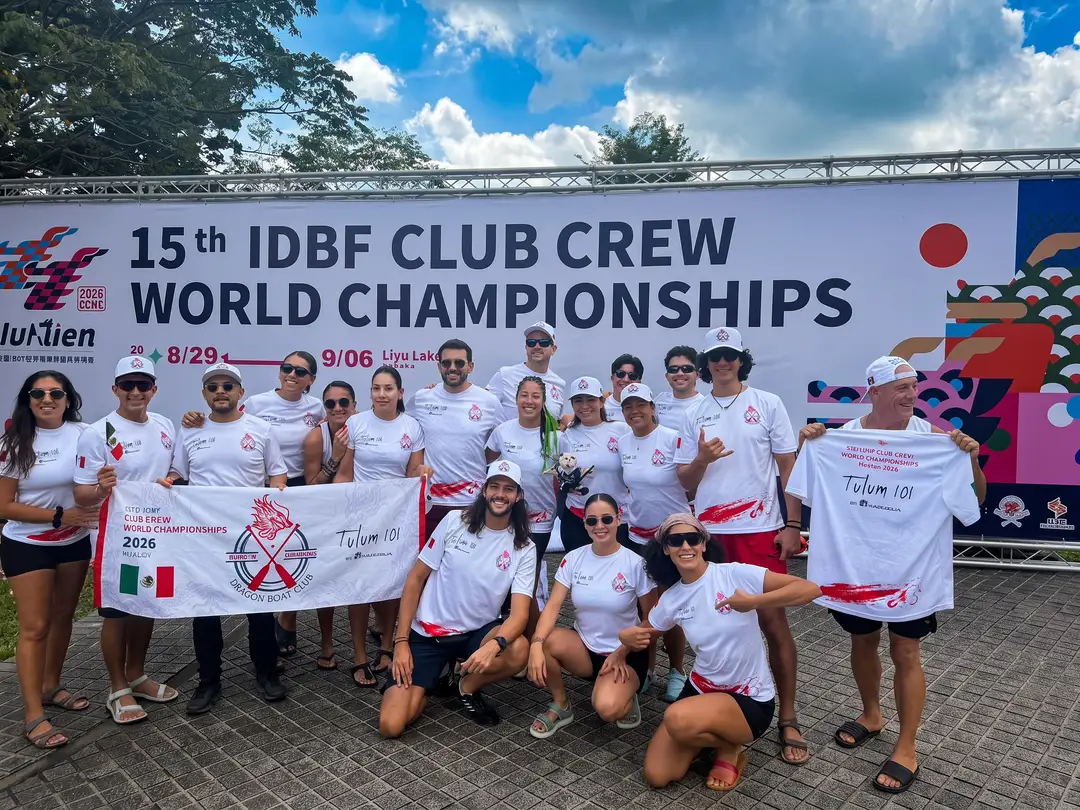 A rowing team in matching white and red uniforms poses together in front of a banner for the 15th IDBF Club Crew World Championships
