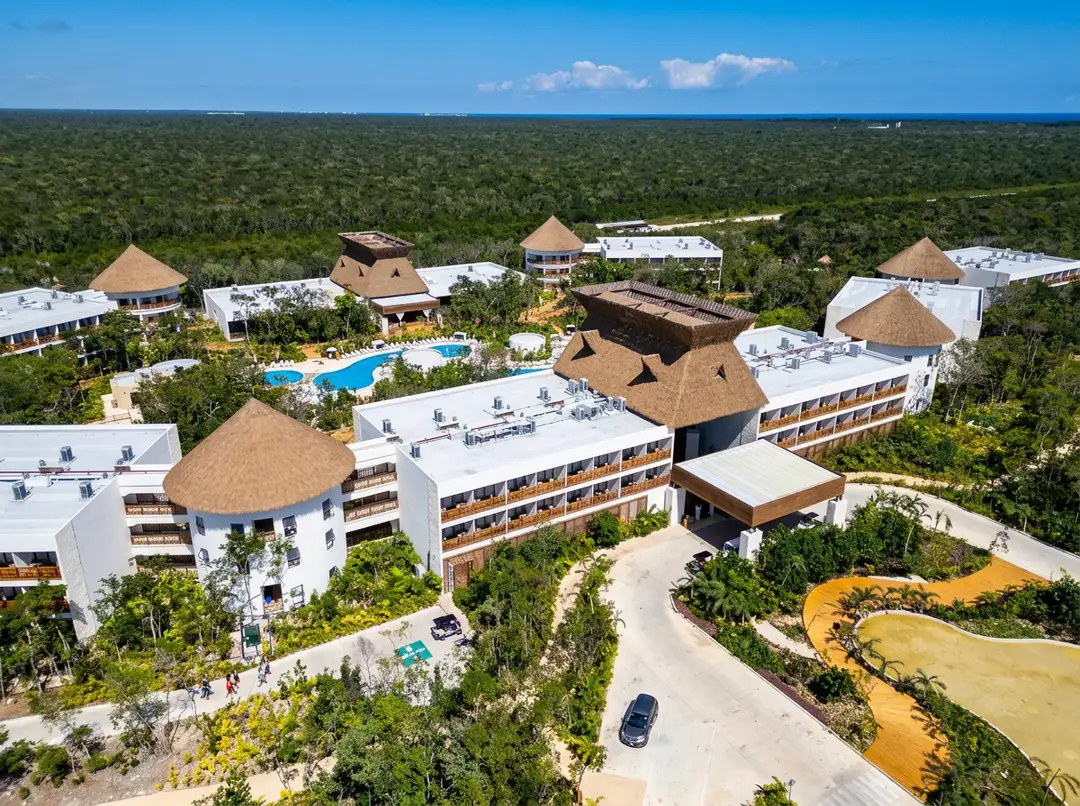 Aerial view of a large resort complex with white buildings featuring thatched roofs, a blue swimming pool, and yellow water slide surrounded by dense jungle for