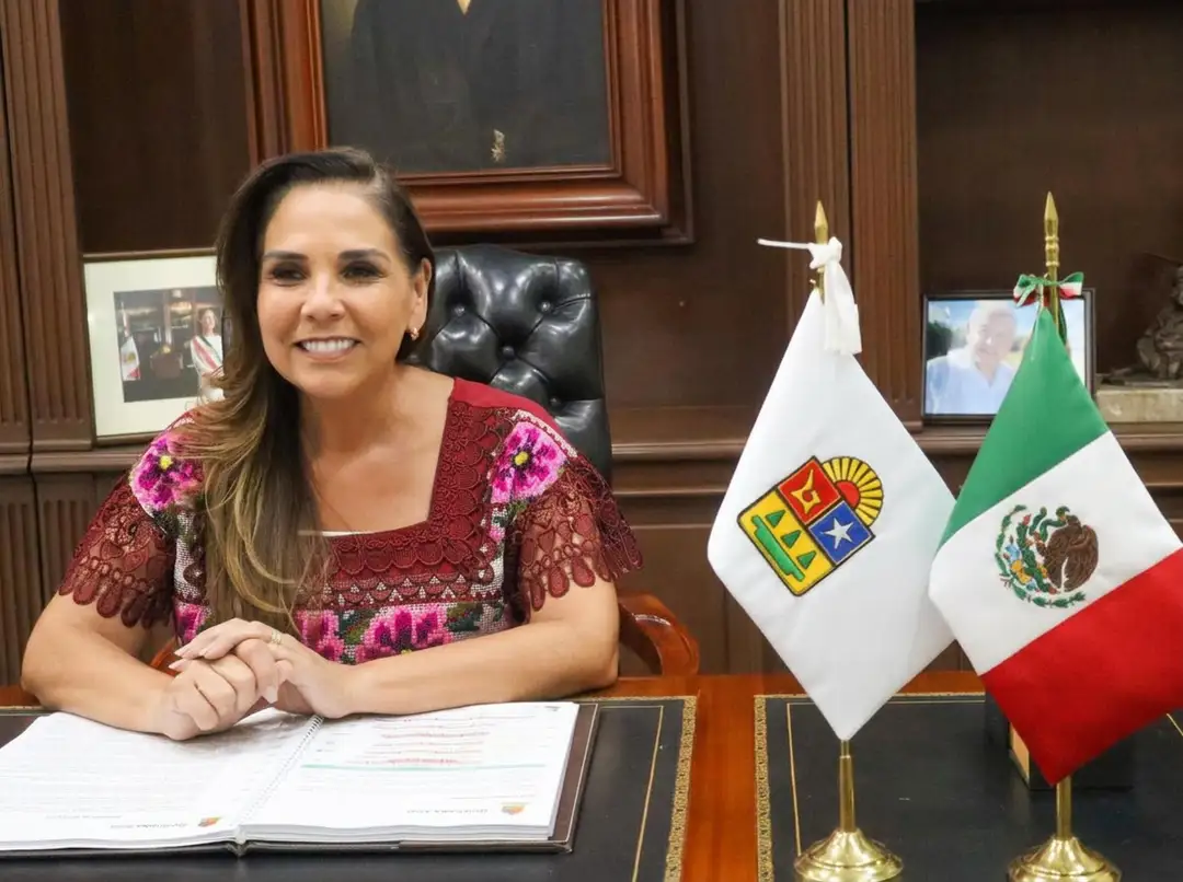 Woman in a burgundy patterned blouse sits at a desk with an open document, with Mexican and regional flags displayed beside her in an official wood-paneled offi