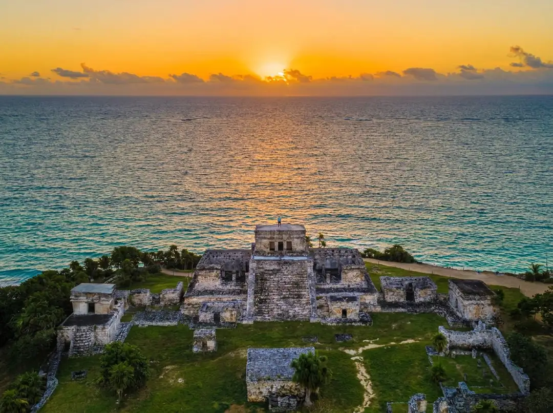 Ruins of a stone pyramid and fortress structure on a grassy coastal promontory overlooking turquoise water at sunrise