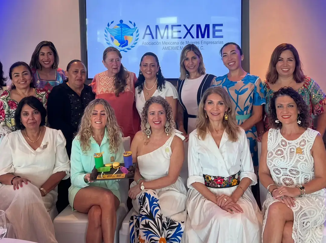 Group of women posing together in front of an AMEXME logo screen at what appears to be a business event or conference