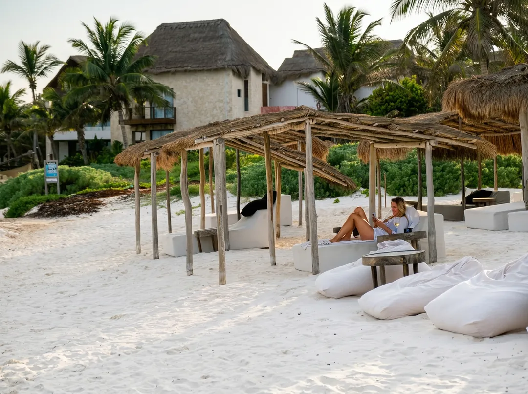 World Cup and Summer Vacations Could Push Tulum Occupancy to 80 Percent? Two people sit under a thatched-roof shelter on a beach with white cushioned seating in the foreground and colonial-style buildings with palm trees in the backg