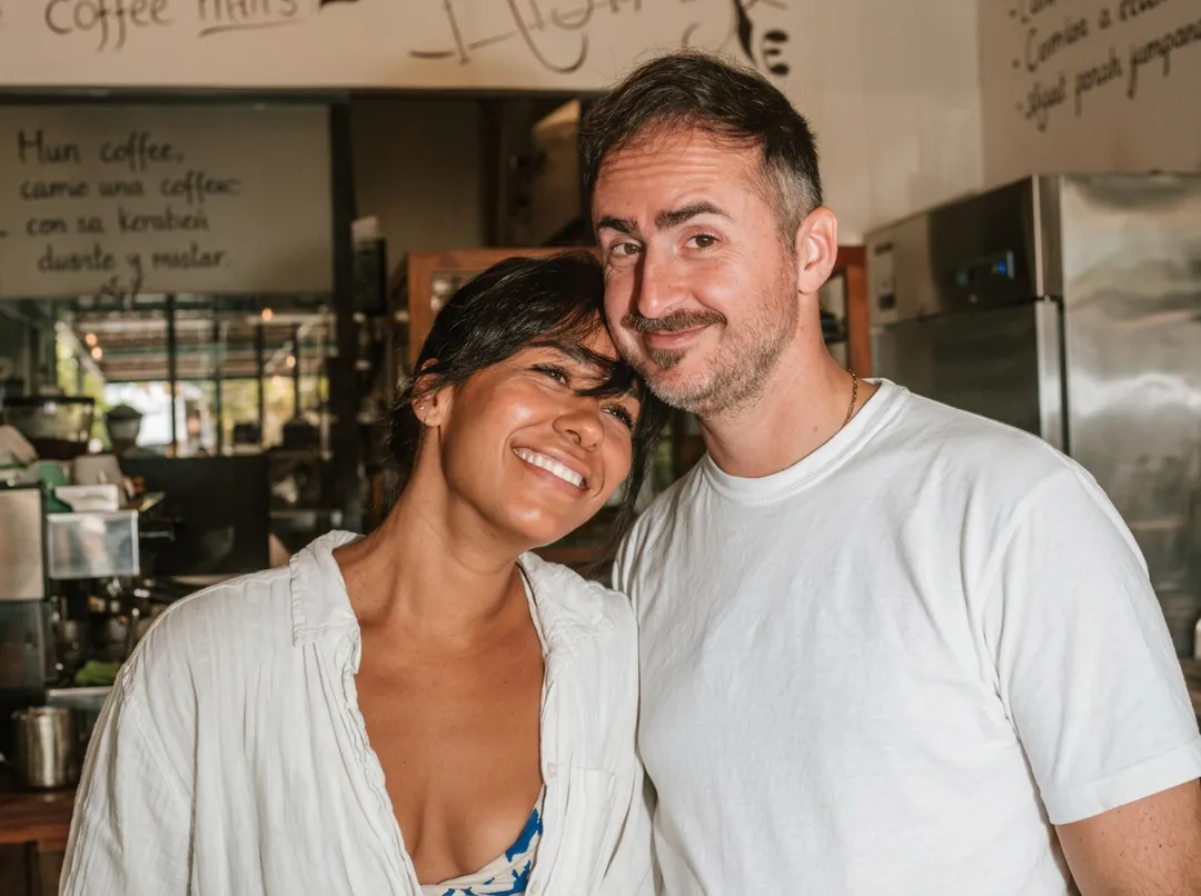 Two people smile together inside a coffee shop with handwritten signs and espresso equipment visible behind them