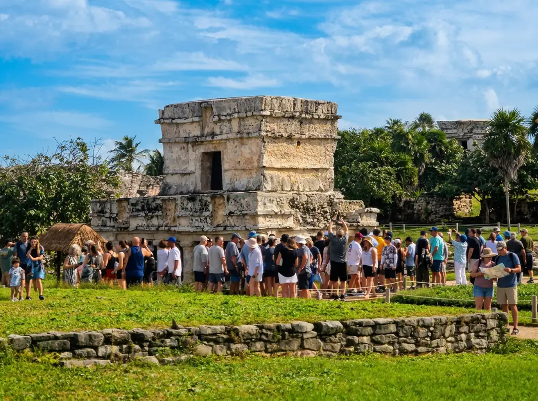 Large crowd of tourists gathered around an ancient stone tower structure surrounded by green grass and tropical vegetation