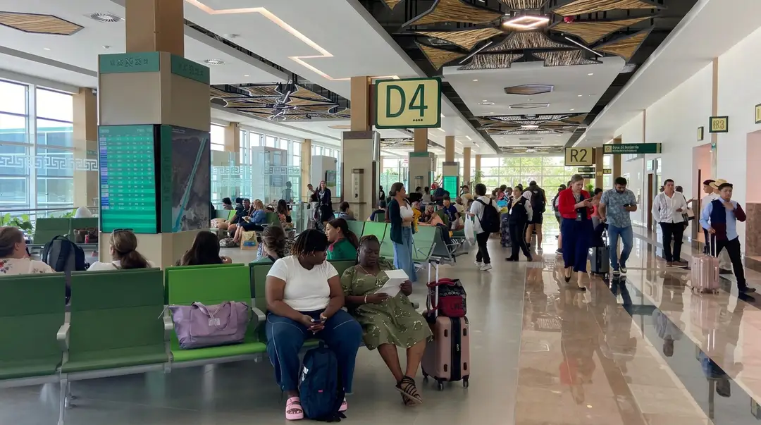 Passengers wait and move through a busy airport terminal with green seating areas and directional signage overhead