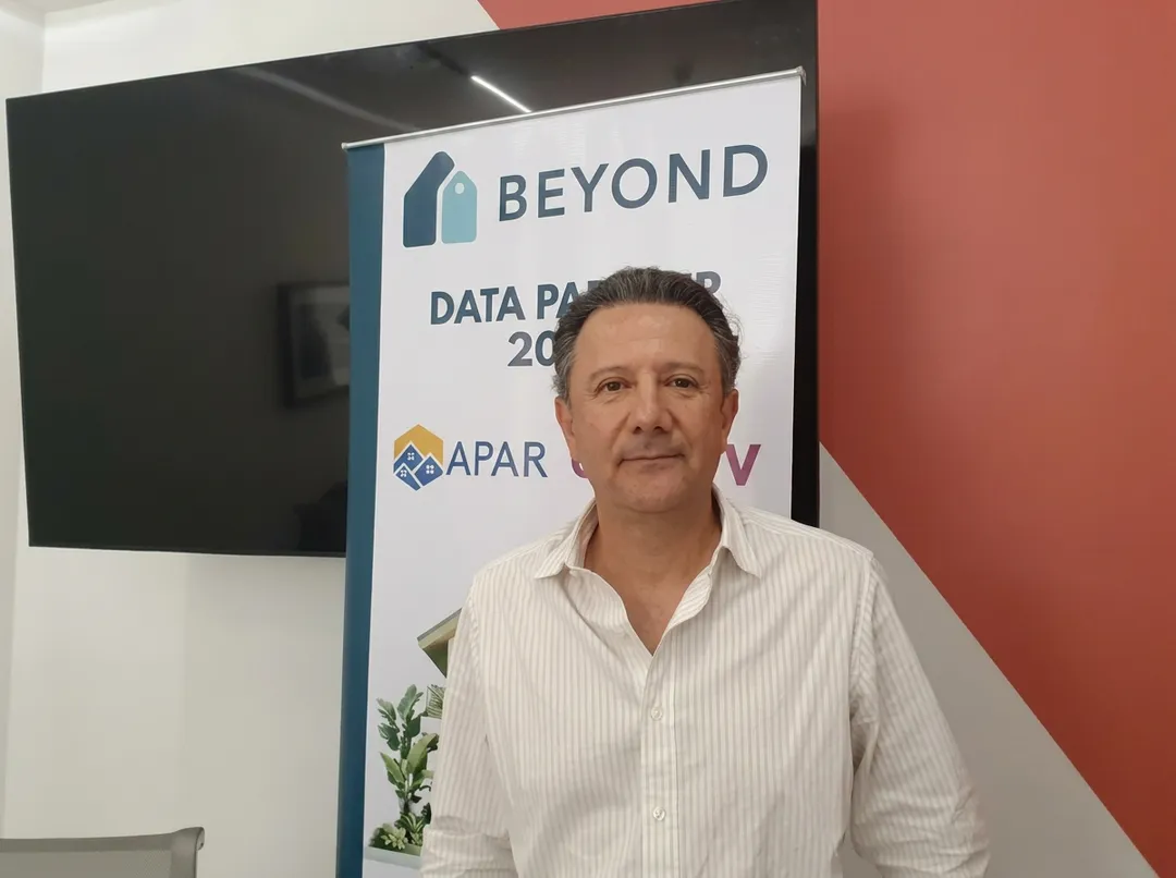 Man in white striped shirt standing in front of a banner with "Beyond" branding and logos at what appears to be a conference or event space