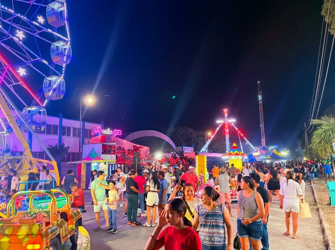 Crowds of people enjoy an evening at an illuminated carnival or fair with colorful rides, including a lit Ferris wheel and other attractions along a beachfront