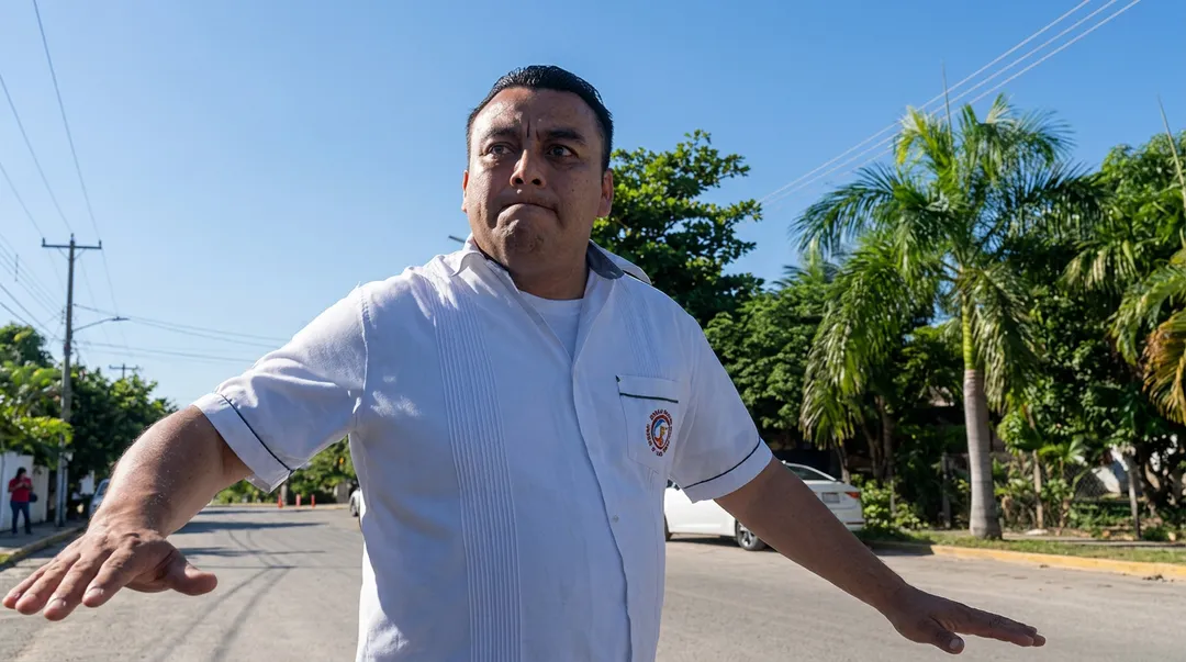 Hombre con camisa blanca de pie en una calle residencial con palmeras y líneas eléctricas al fondo.
