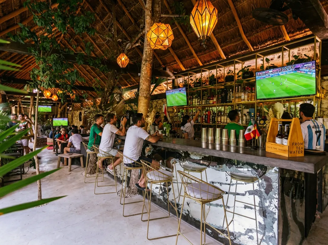 Patrons sit at a bar watching soccer matches on multiple screens in an open-air venue with wooden beams and hanging lanterns