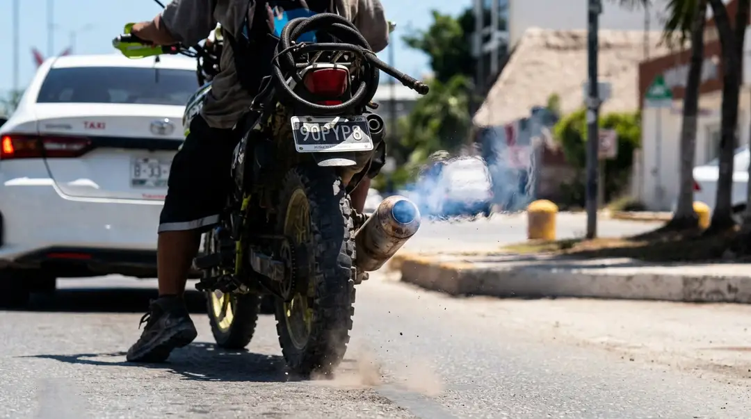 A motorcycle with a modified exhaust pipe emits smoke while parked on a city street near a taxi and palm trees