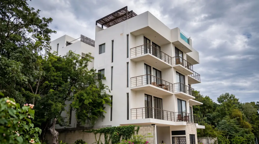 4,500 vacation rentals in Tulum, and no map of the ones inside irregular settlements Modern white apartment building with multiple balconies surrounded by green vegetation and trees under a cloudy sky
