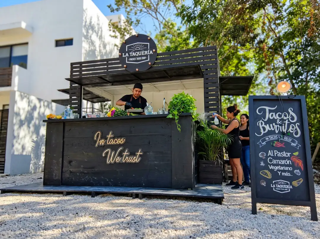La Veleta goes first as Tulum's public space operation targets workshops and street vendors A black food cart vendor serves customers from a window counter in an outdoor public space, with a menu board and planted greenery visible nearby