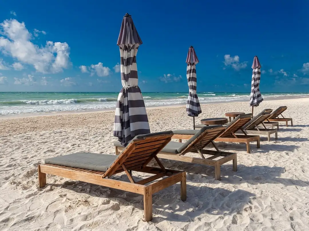Wooden lounge chairs with gray cushions and striped umbrellas line a sandy beach with turquoise water and waves under a blue sky