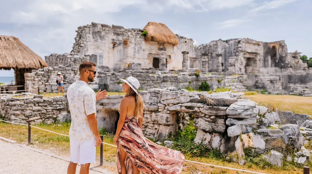 Two tourists stand on a path viewing ancient stone ruins with thatched structures nearby