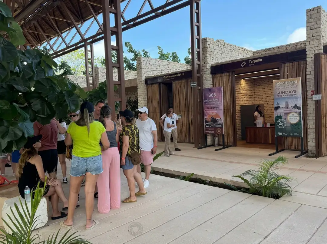 Visitors queue outside a wooden-fronted entrance building at an archaeological site visitor area with stone walls and metal framework overhead
