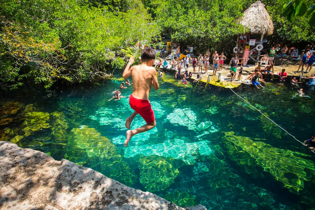 Facing Another Sargassum Season, Tulum Bets on Cenotes to Keep Visitors Coming Shirtless man jumps into clear turquoise cenote water surrounded by swimmers and beachgoers near a thatched-roof structure