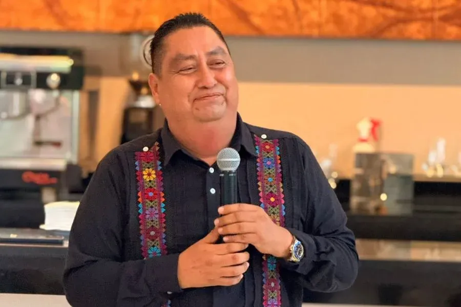 Man wearing a dark shirt with colorful embroidered trim holds a microphone while speaking indoors
