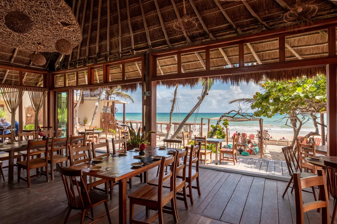 Eight to Fifteen Restaurants Close in Tulum as Sector Reports Worst Season in Years Wooden-framed beachfront restaurant interior with dining tables overlooking the ocean and beach where visitors are seated