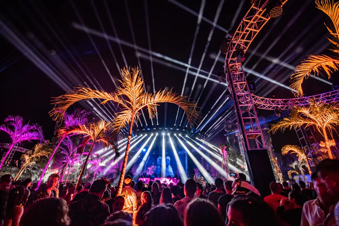 Crowd of people at an outdoor music festival with illuminated palm trees, a roller coaster structure, and blue stage lights beaming into the night sky