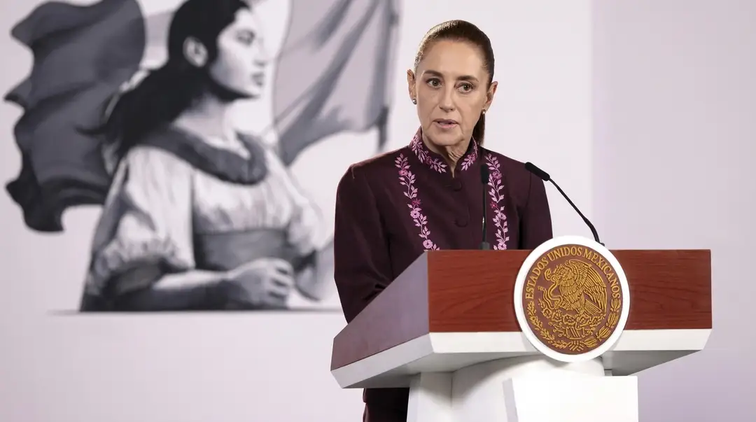 Sheinbaum responde a la pregunta de seguridad detrás de los vuelos a Tulum Woman in maroon embroidered shirt speaking at a podium with an official seal, with a mural of a figure visible in the background