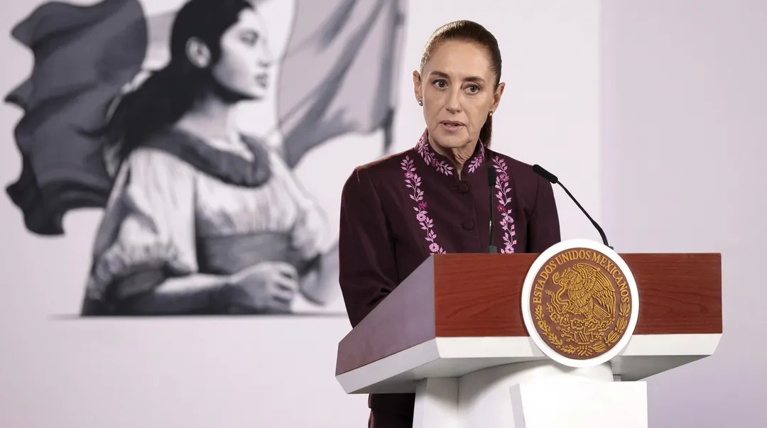 Woman in maroon embroidered shirt speaking at a podium with an official seal, with a mural of a figure visible in the background