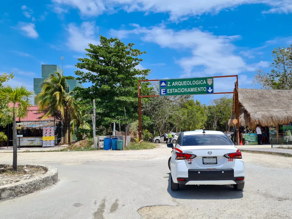A white sedan drives toward a directional sign for an archaeological site and parking area, with palm trees and thatched-roof structures visible along the road