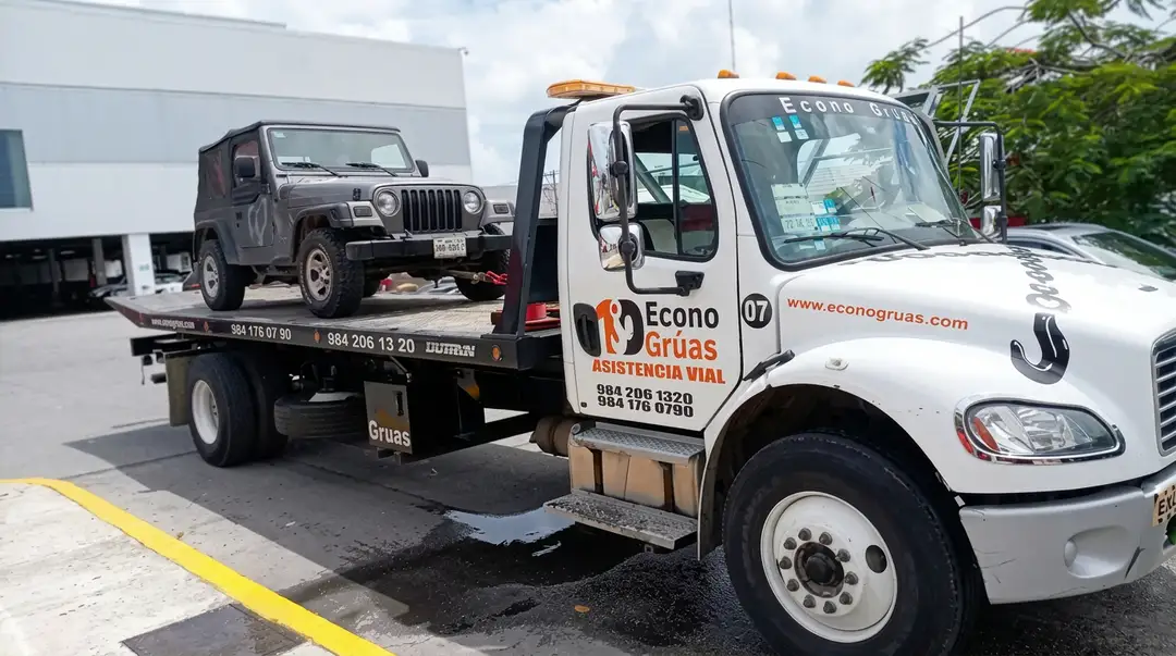 A white tow truck carries a dark-colored jeep on its flatbed in a parking lot