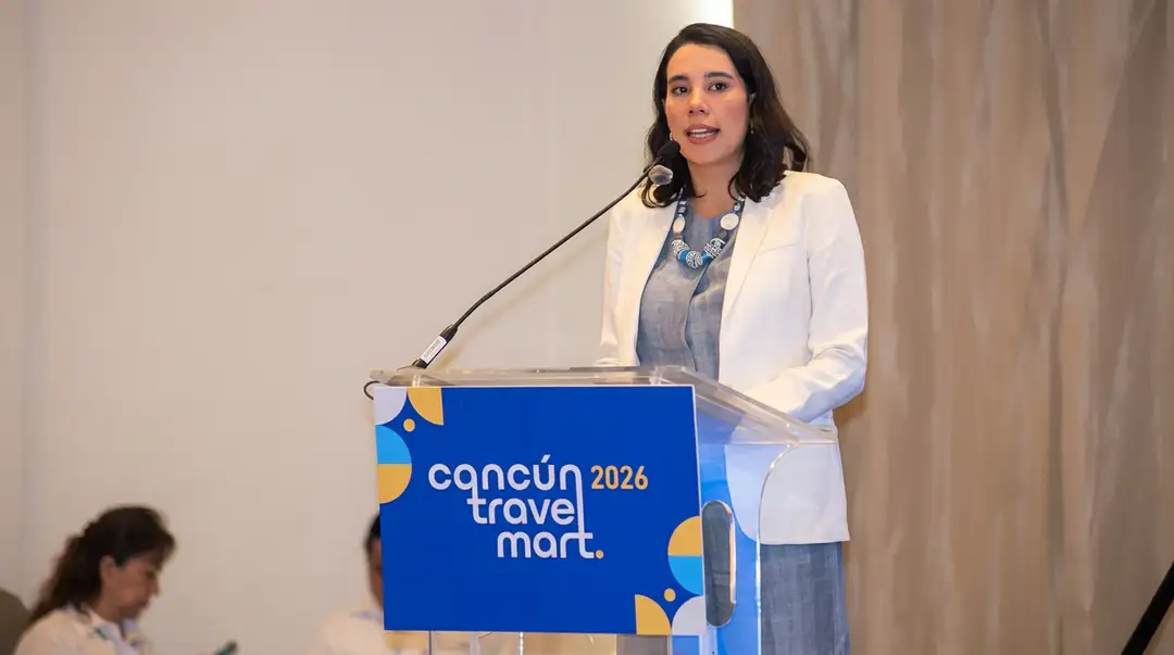 Woman in white blazer speaking at a podium with a microphone during a travel industry event