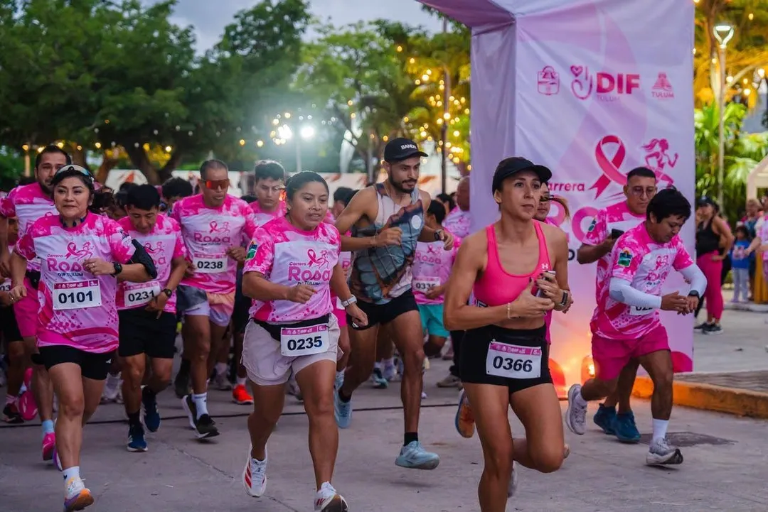 Participants wearing pink race bibs and shirts run through a street at night past a large pink banner with breast cancer awareness symbols