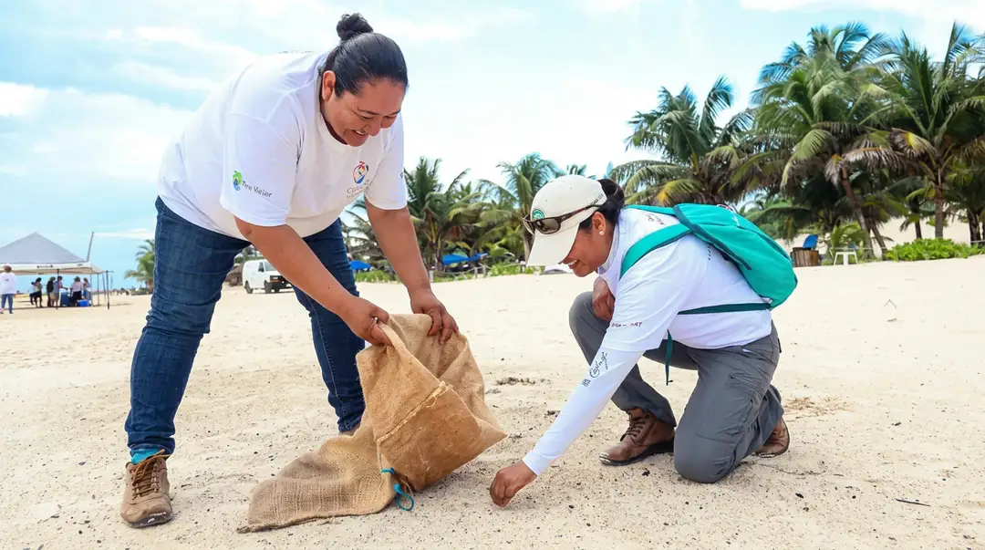 Tulum’s Conservation Week Opens Its Weekend Two people collect trash in a burlap sack on a sandy beach lined with palm trees